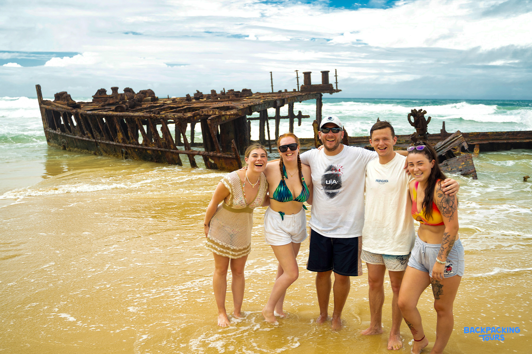 Five happy backpackers pose in front of the historic Maheno shipwreck on Fraser Island, Australia.