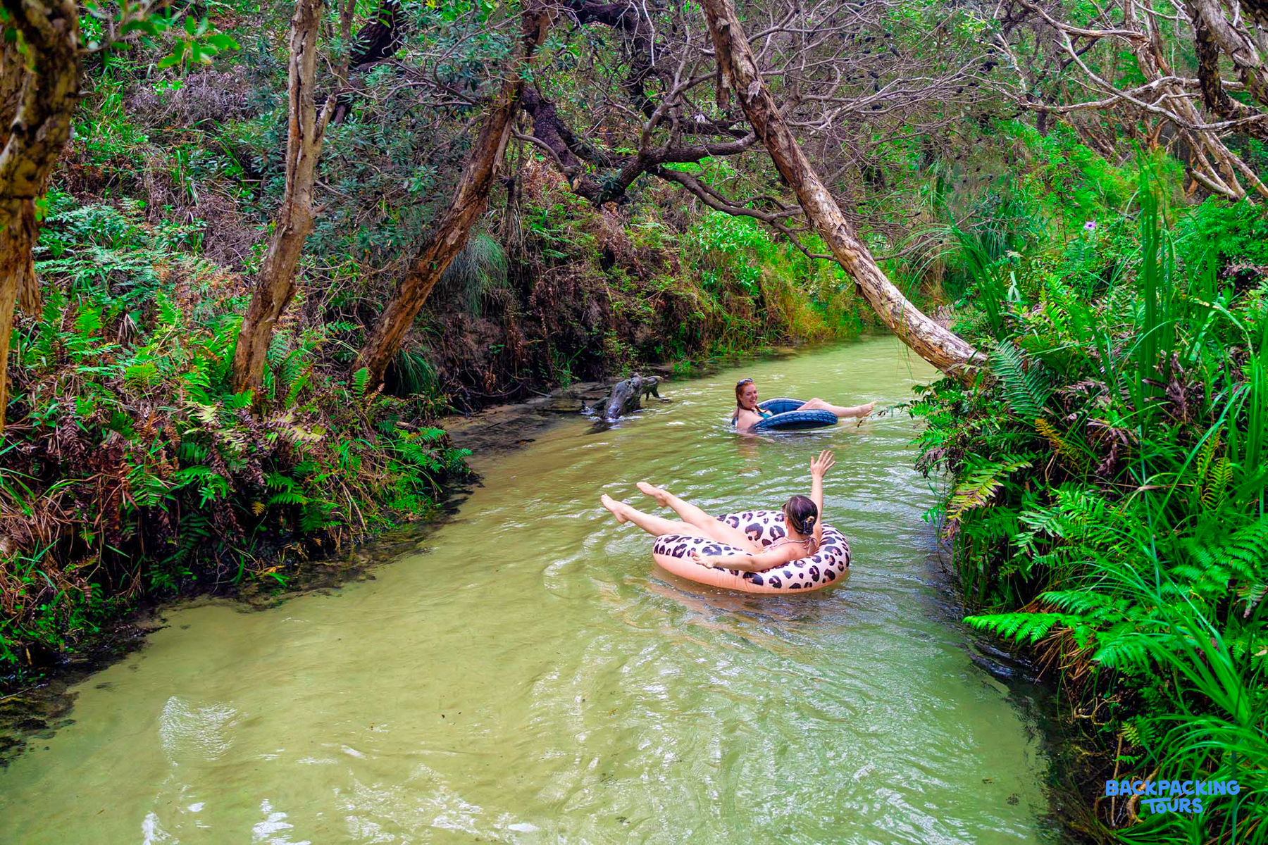 Two people happily float down Eli Creek on K'gari, surrounded by lush green foliage.