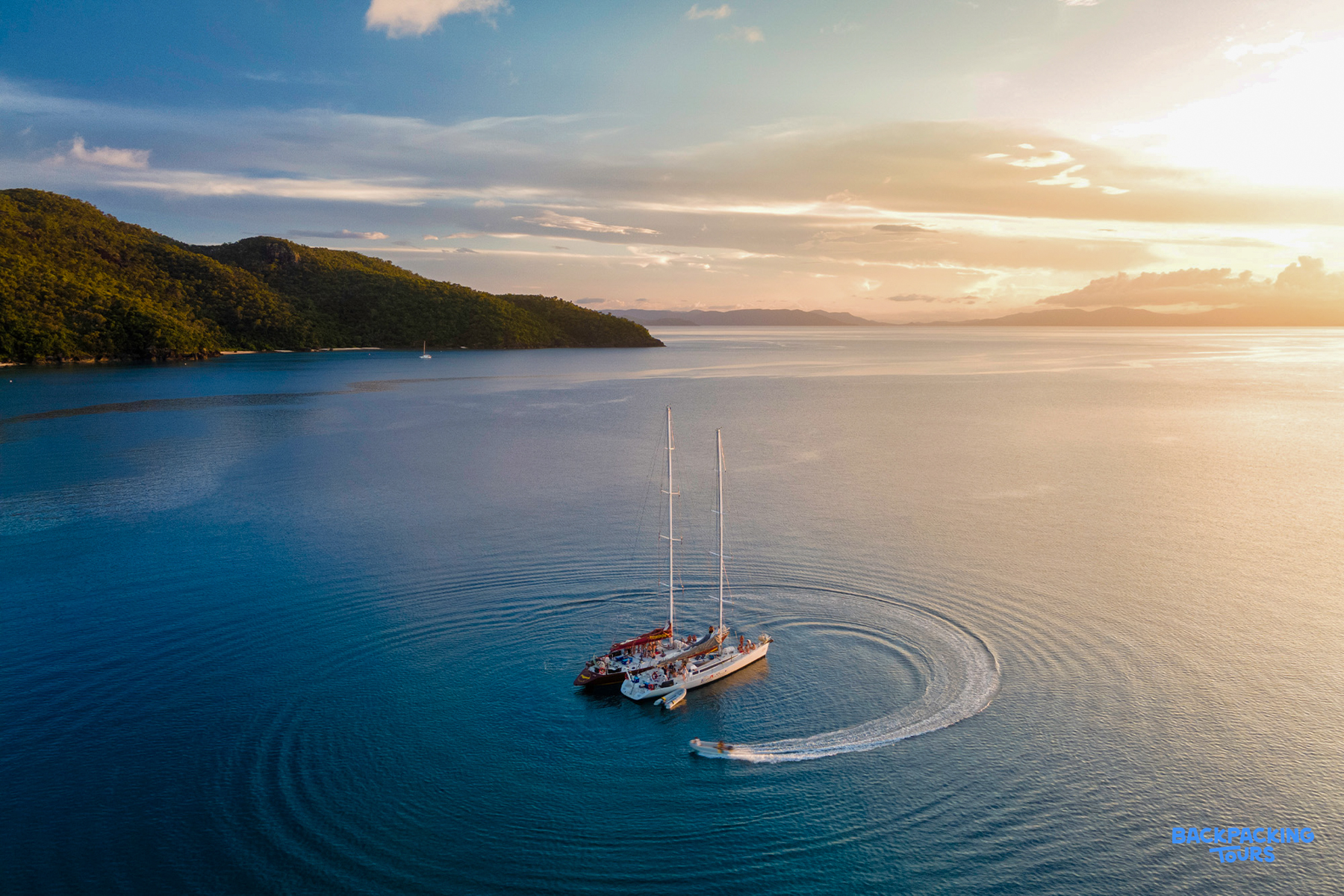 A large catamaran and small boat make ripples in the calm blue Whitsundays waters at sunset.