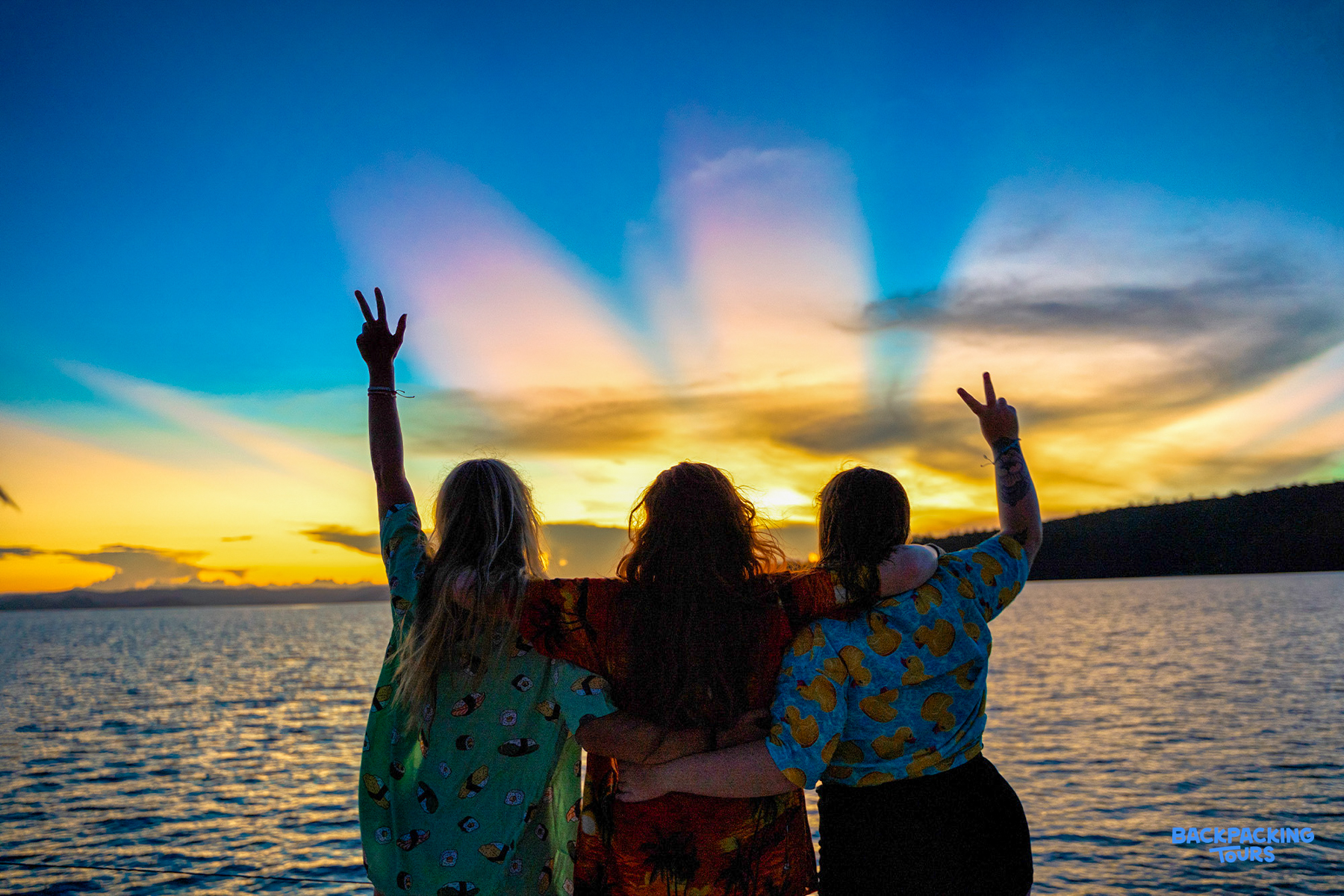 Three friends embrace, making peace signs at a beautiful Whitsundays sunset.