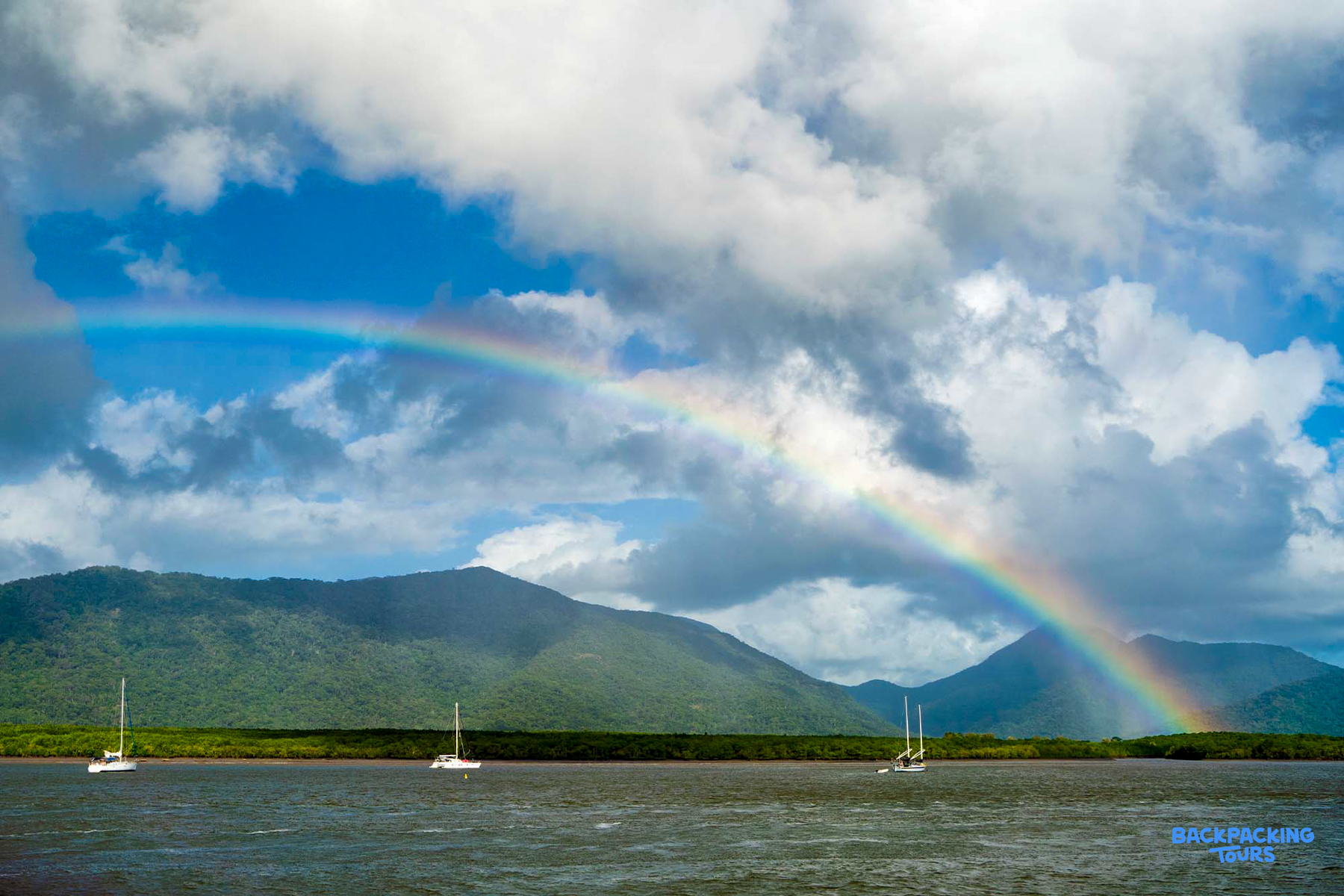 A vibrant rainbow arches over a bay with sailboats and lush green mountains in Cairns.