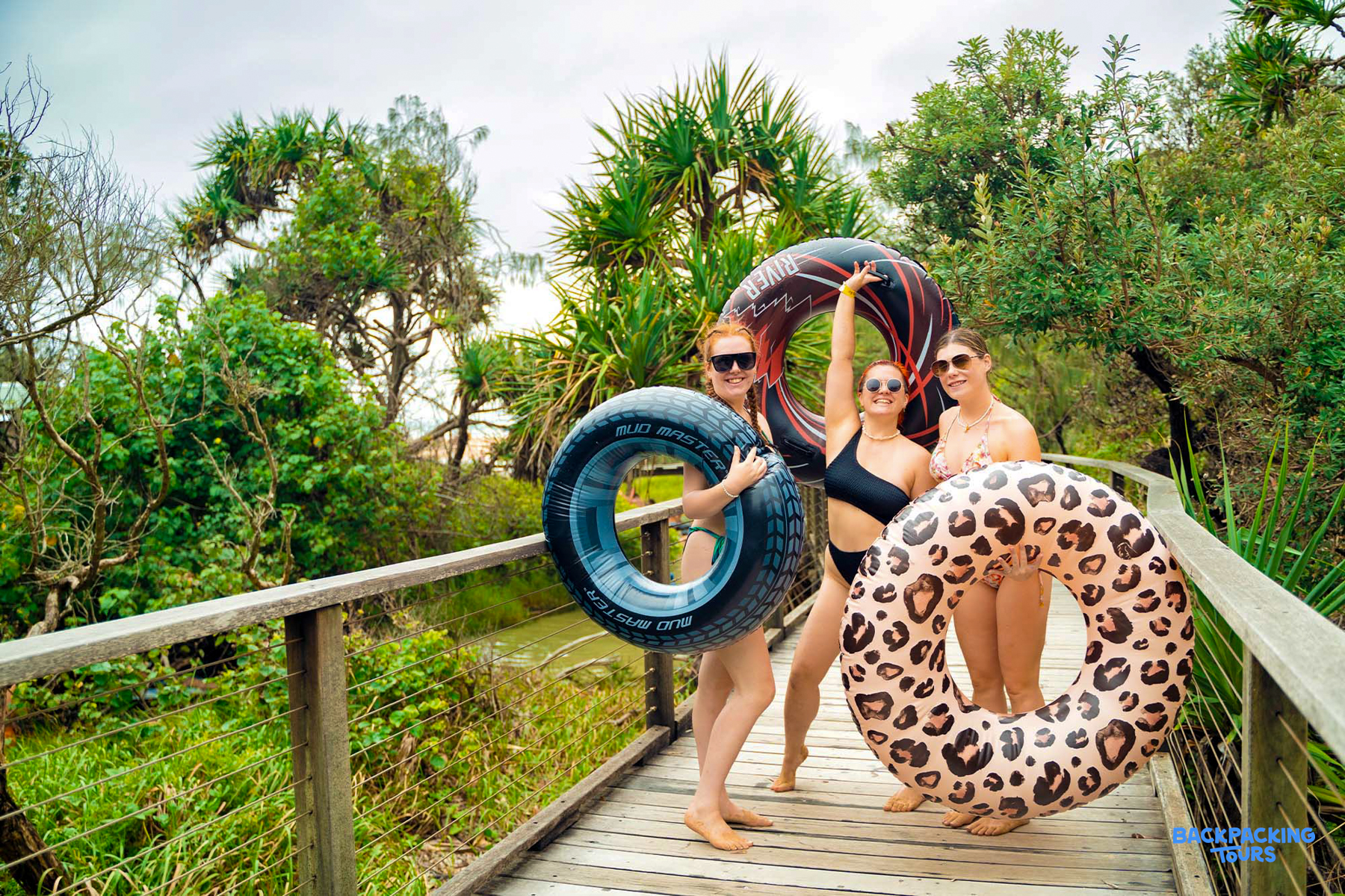 Three cheerful women on a wooden boardwalk, holding inflatable tubes, ready for a tropical water adventure.
