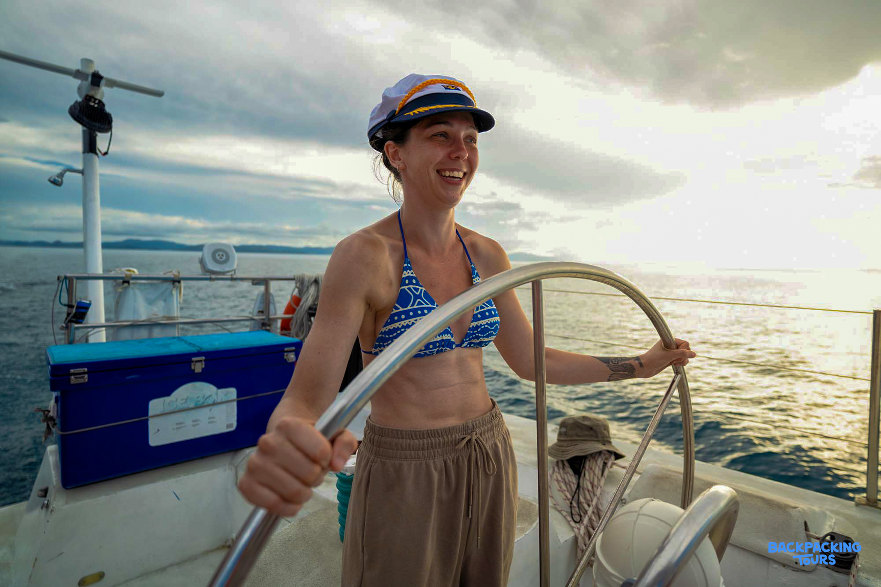 A smiling woman in a captain's hat steers a boat through the Whitsundays at sunset.