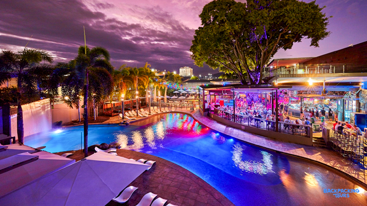 Lively pool area at Gilligan's Hostel in Cairns, illuminated by colorful lights under a dramatic purple sky.