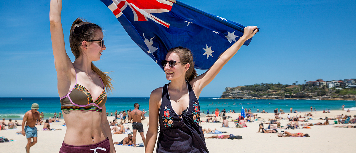 Two smiling women joyfully wave an Australian flag on a crowded, sunny Bondi Beach.