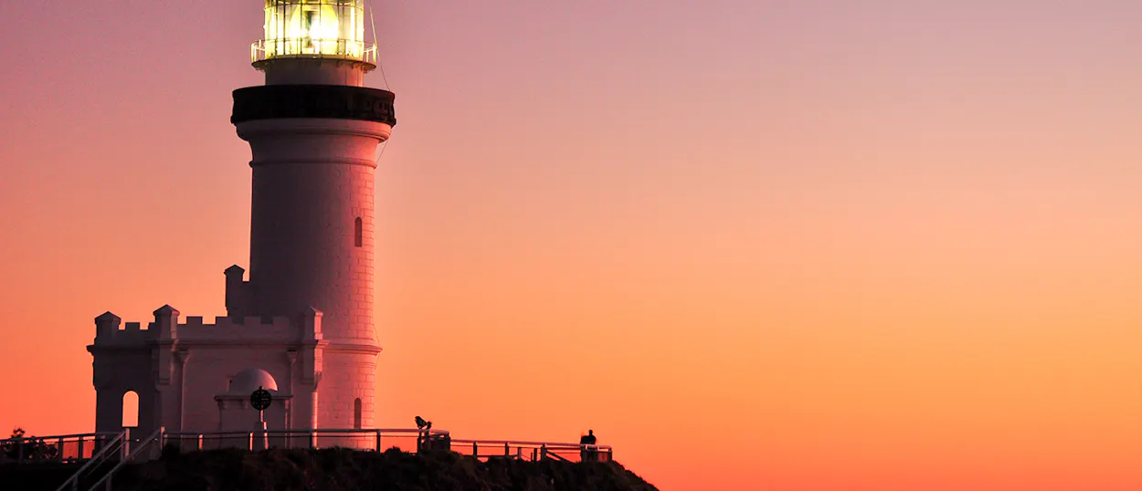 Cape Byron Lighthouse stands illuminated against a stunning orange and pink sunrise sky over the ocean.
