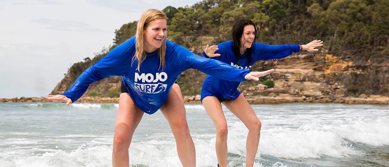 Two smiling women in blue surf shirts balancing on boards during a lesson in the ocean.