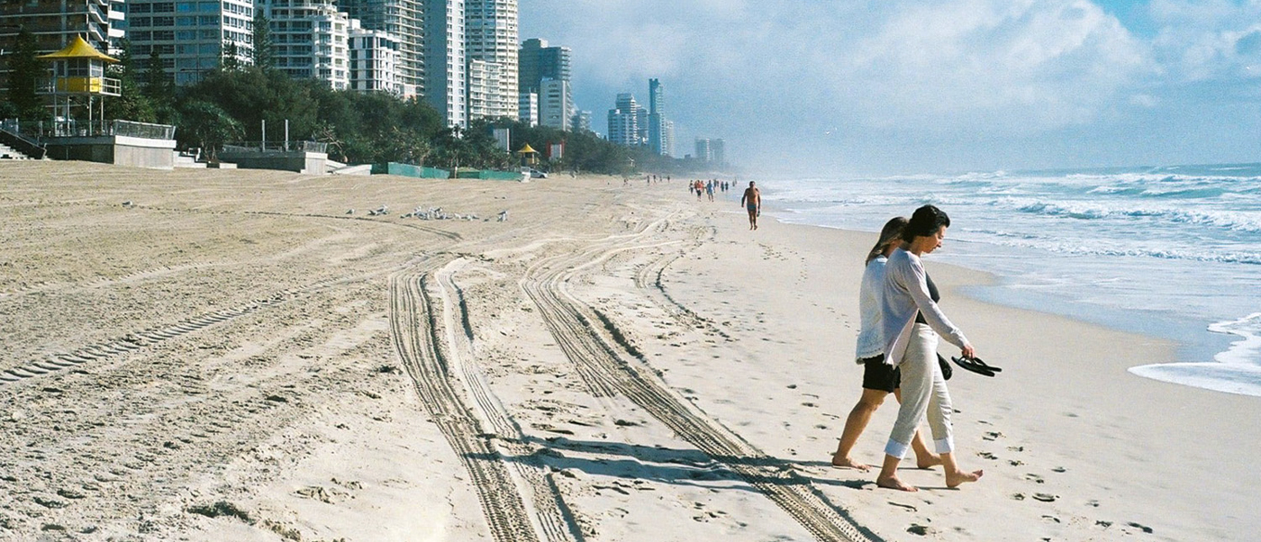 Barefoot travelers stroll along the golden sands of Surfers Paradise beach with a city skyline behind them.