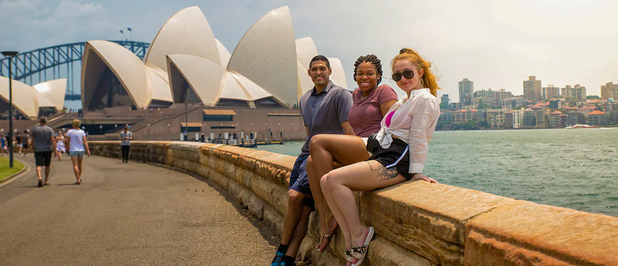 Three smiling friends sit on a wall with Sydney Opera House and Harbour Bridge in the background.