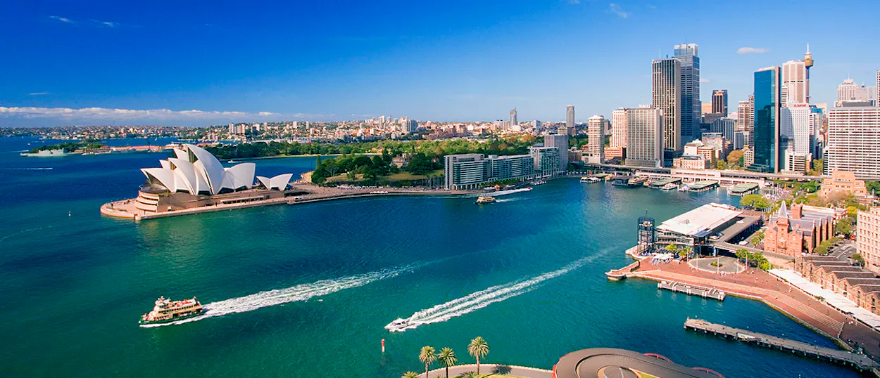 Panoramic view of Sydney Harbour with the iconic Opera House, city skyline, and boats on blue water.