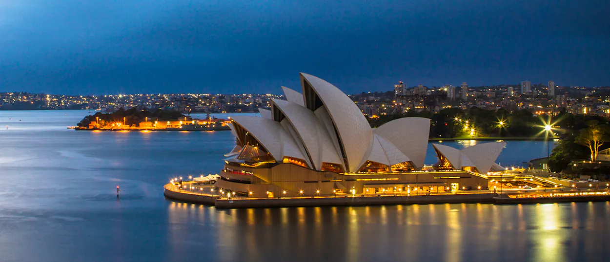 The iconic Sydney Opera House illuminated at dusk, reflecting on the calm harbor waters.