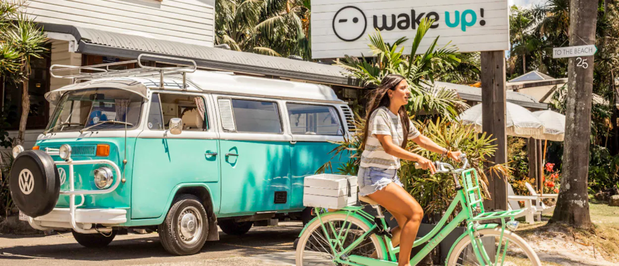 A woman rides a green bicycle past a vintage turquoise van and "Wake Up!" hostel in Byron Bay.