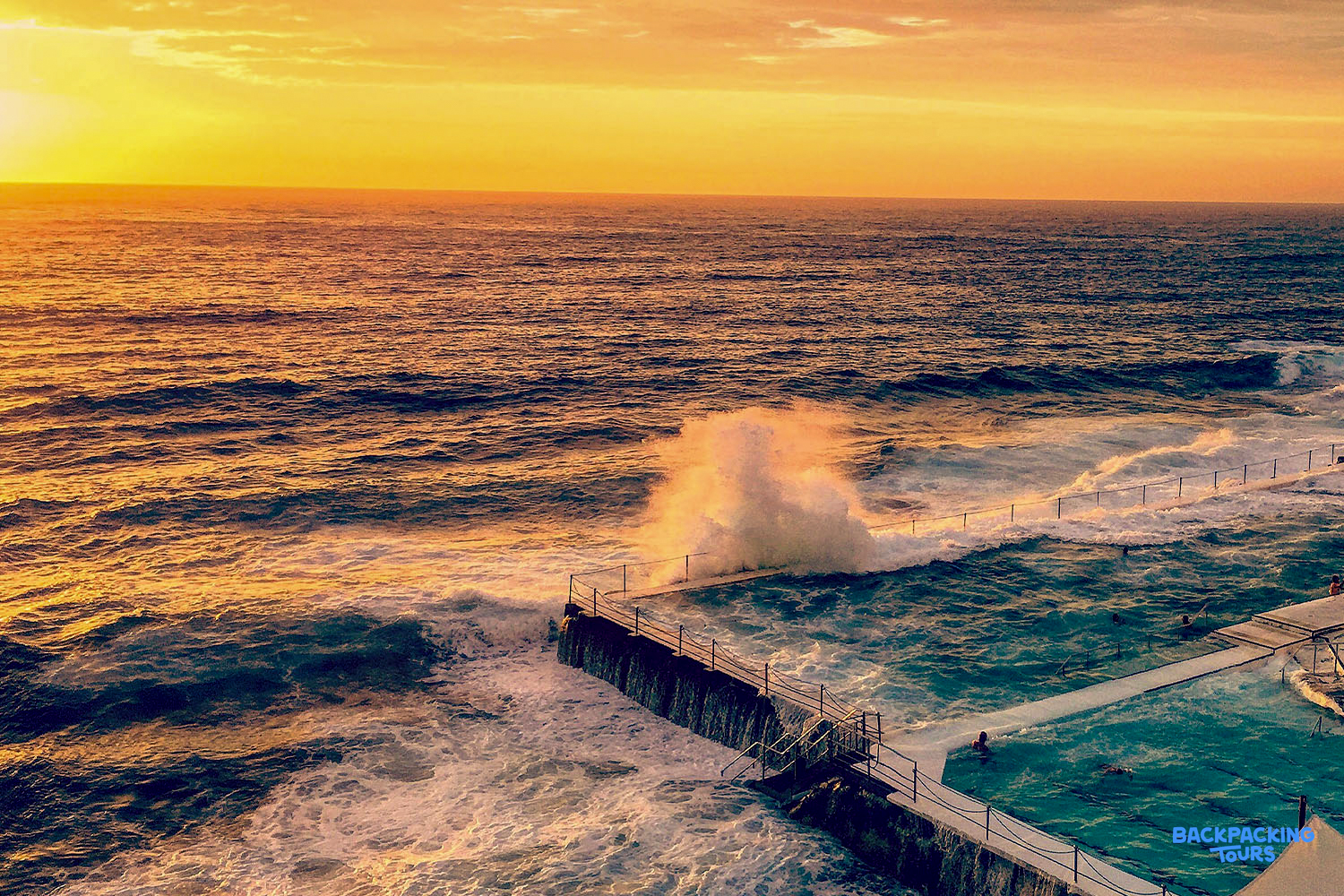 A powerful wave crashes into the ocean wall of Bondi Beach pools under a golden sunset.