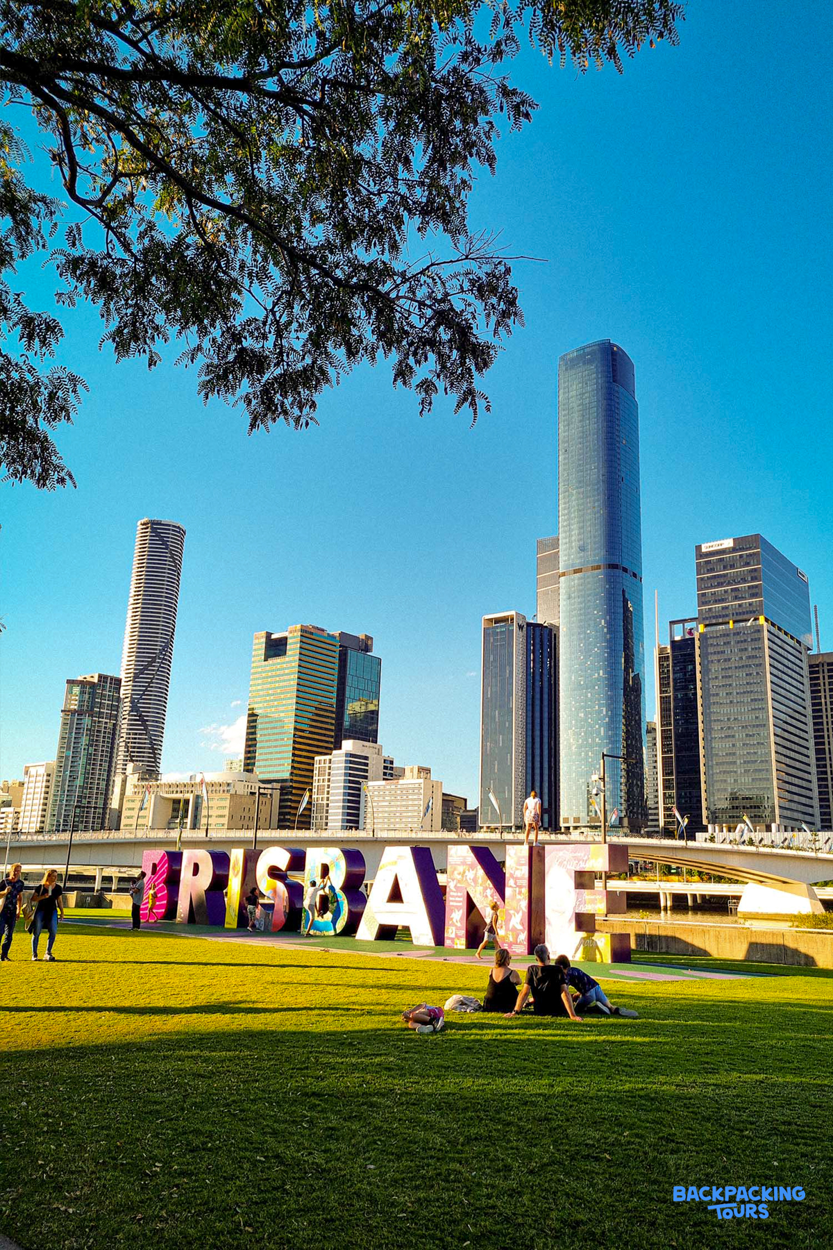 Tourists relax on the grass at Brisbane's South Bank, with the colorful city sign and skyline.