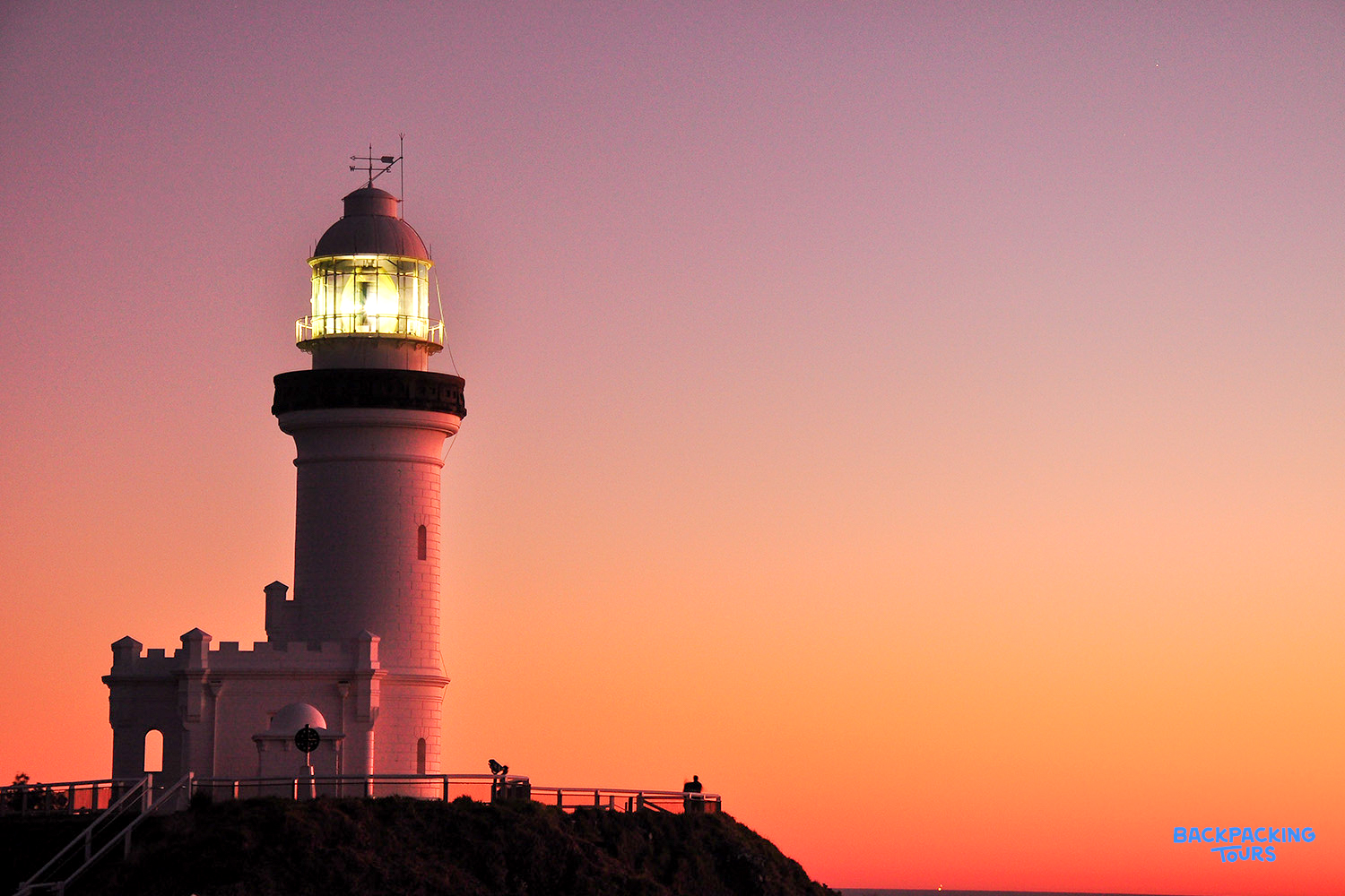 The iconic Cape Byron Lighthouse shines against a vibrant orange and pink sunrise sky.