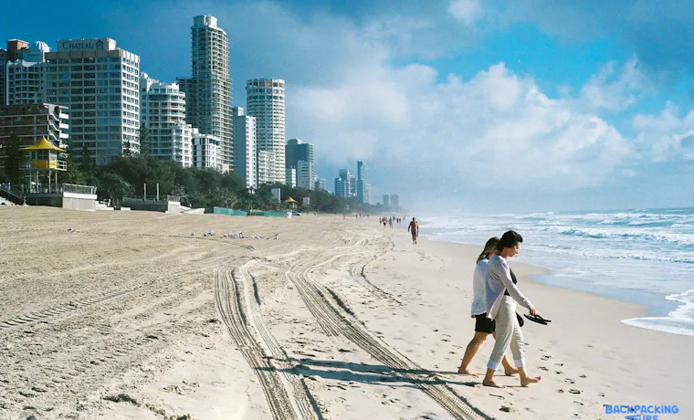 Two people walk barefoot along the golden sands of Surfers Paradise beach, with city skyscrapers in the background.