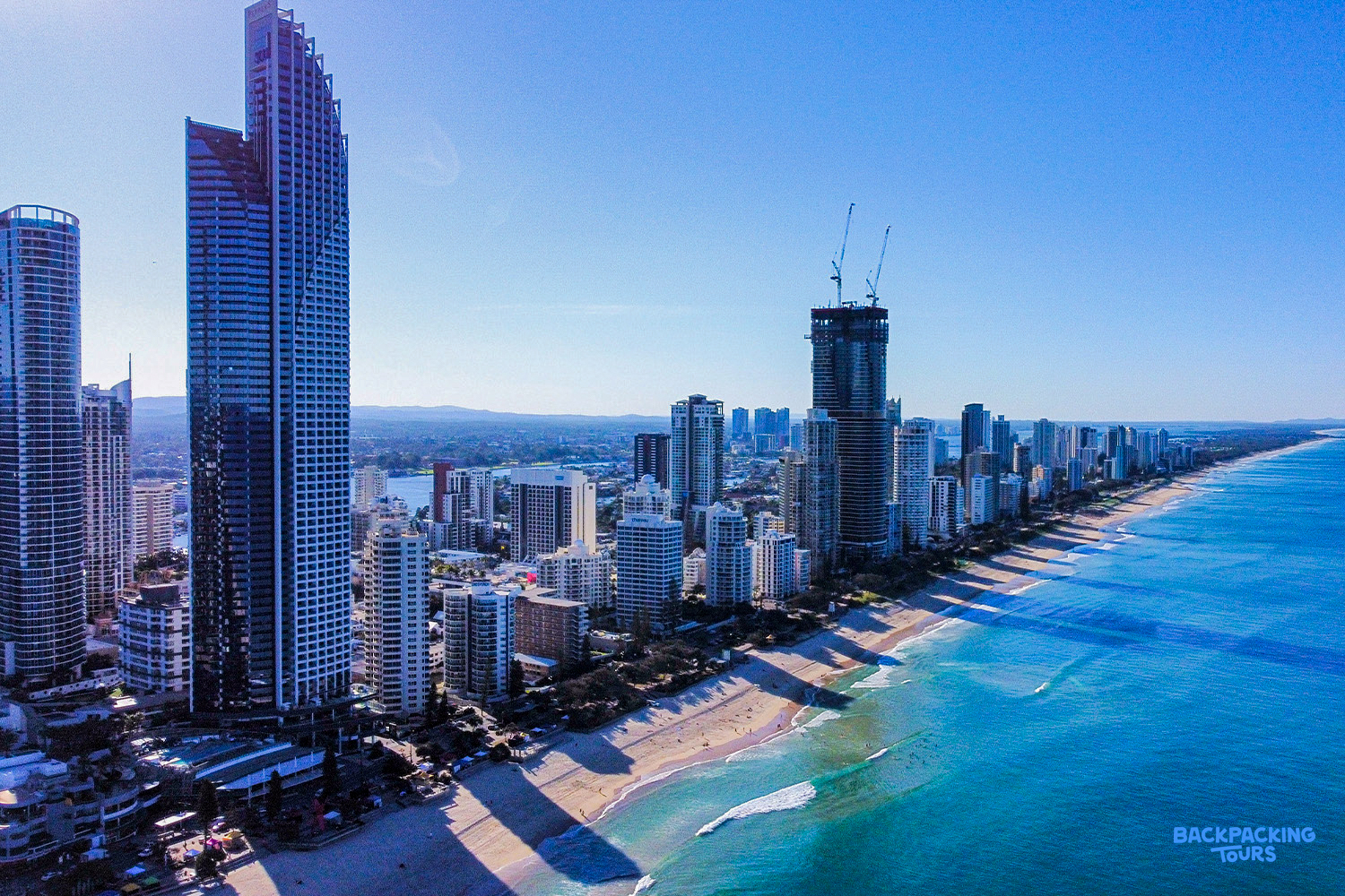 Aerial view of Surfers Paradise skyline with tall buildings lining the long sandy beach and blue ocean.