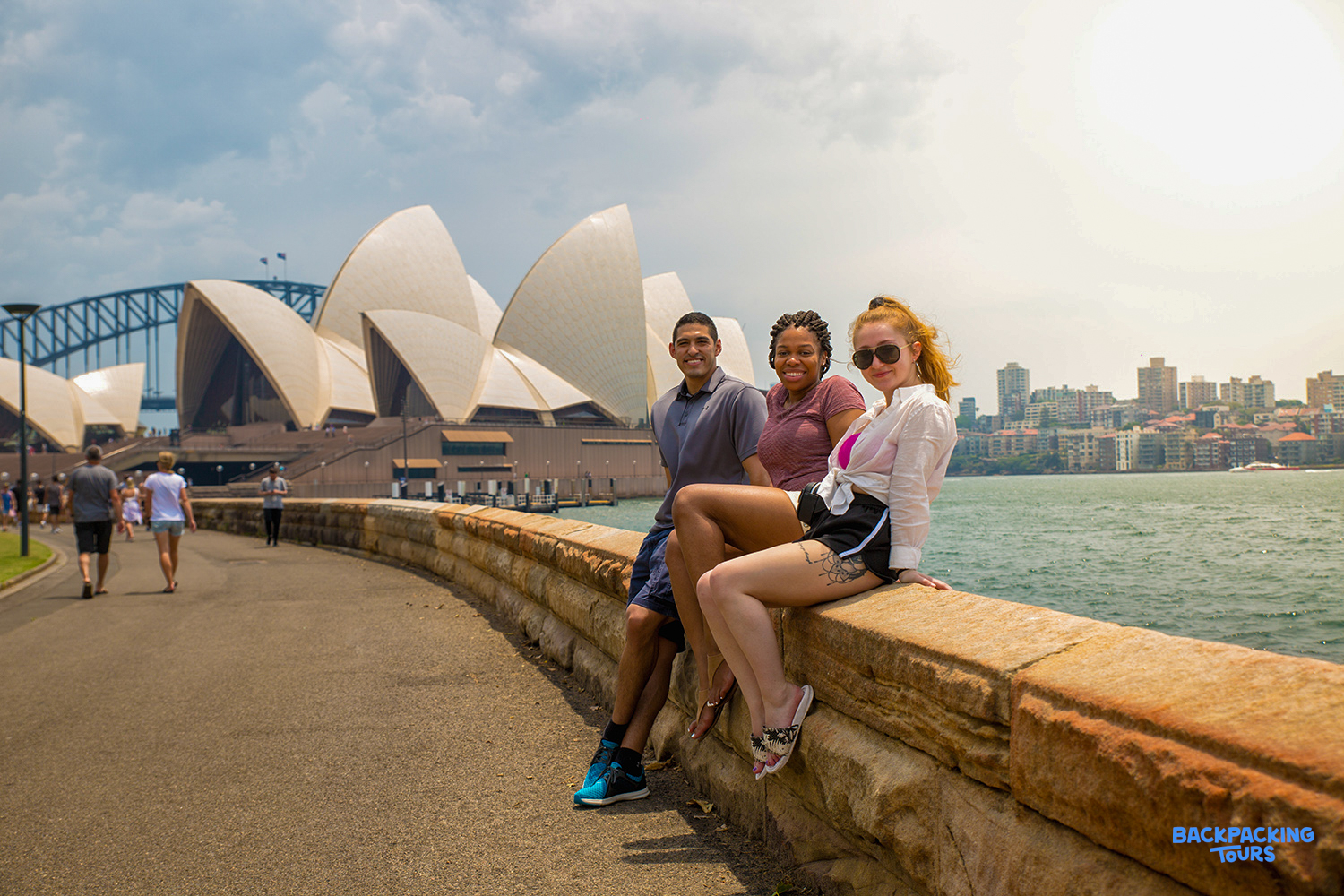 Three happy backpackers pose on a wall with the iconic Sydney Opera House and Harbour Bridge.
