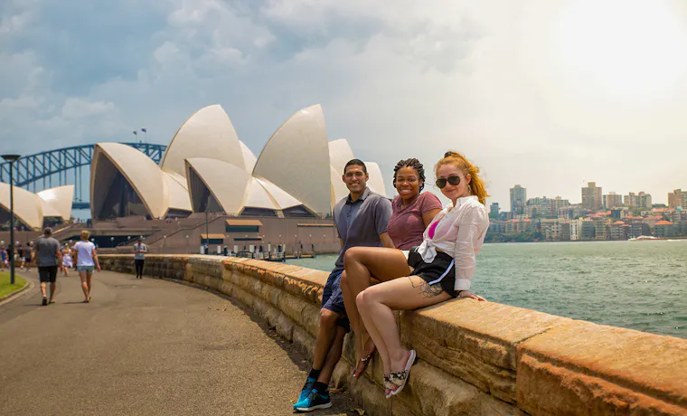 Three happy backpackers pose on a wall with the iconic Sydney Opera House and Harbour Bridge.