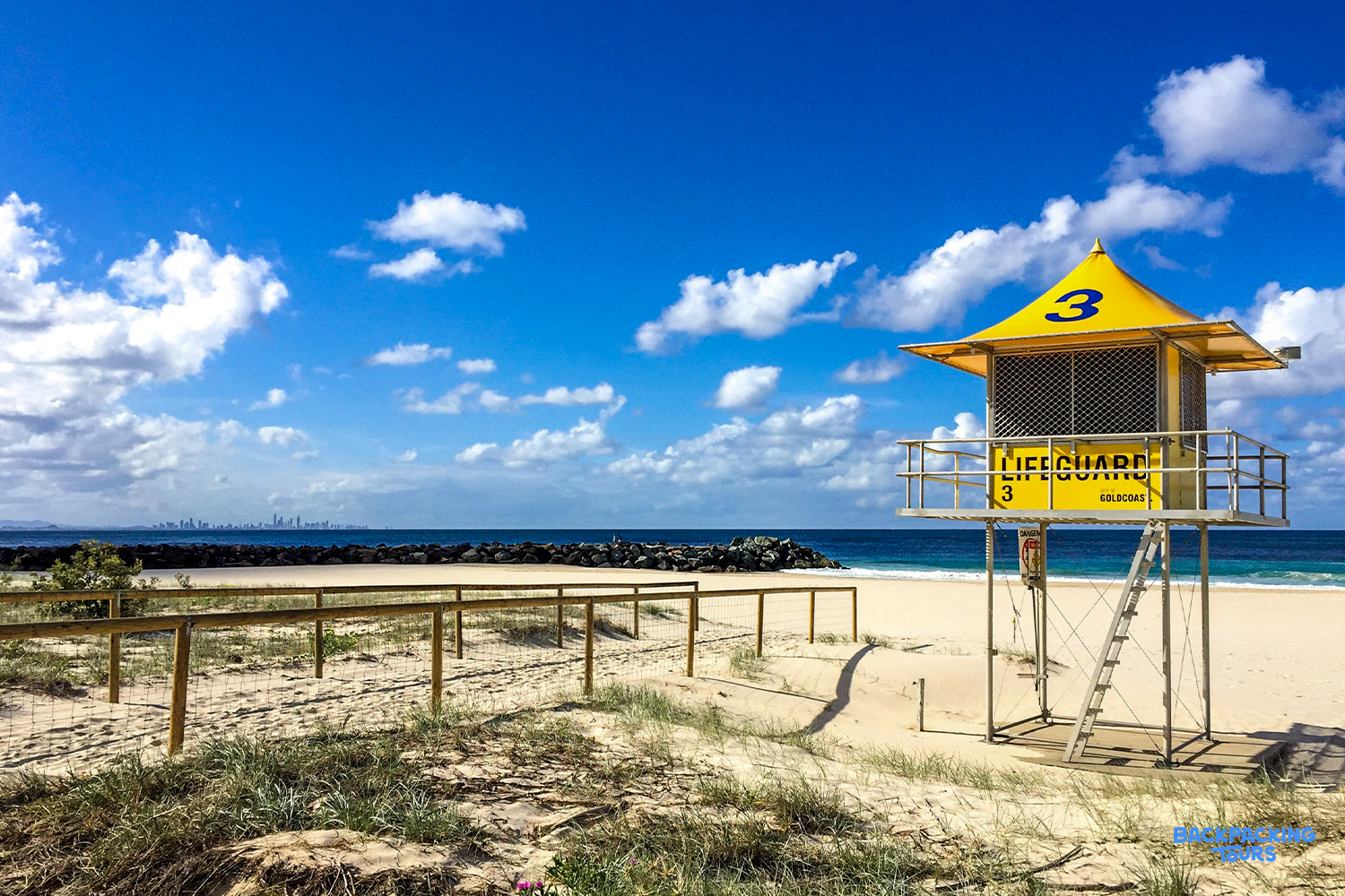 A bright yellow lifeguard tower stands on the golden sands of Surfers Paradise beach under a blue sky.