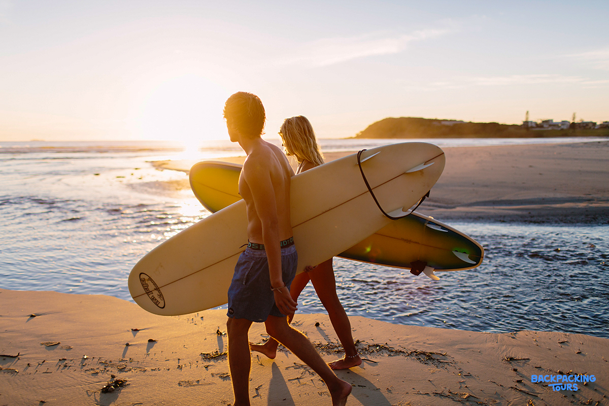 Two surfers walk along the sandy Byron Bay beach at sunset, carrying their boards.