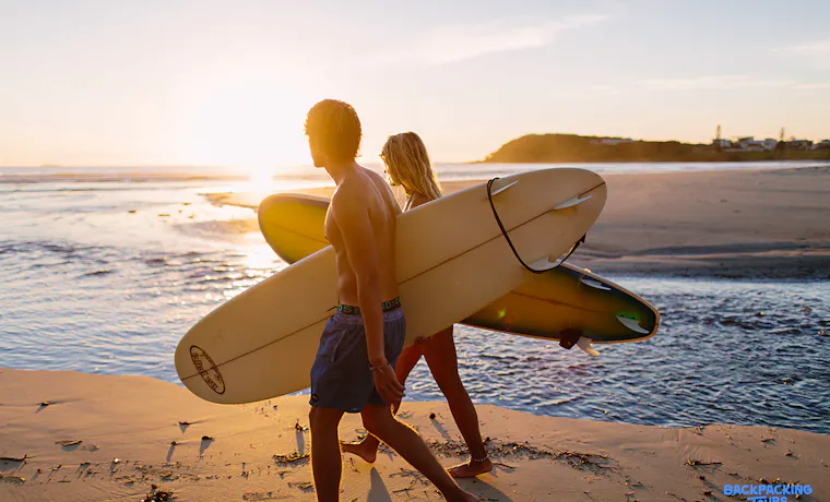 Two surfers walk along the sandy Byron Bay beach at sunset, carrying their boards.