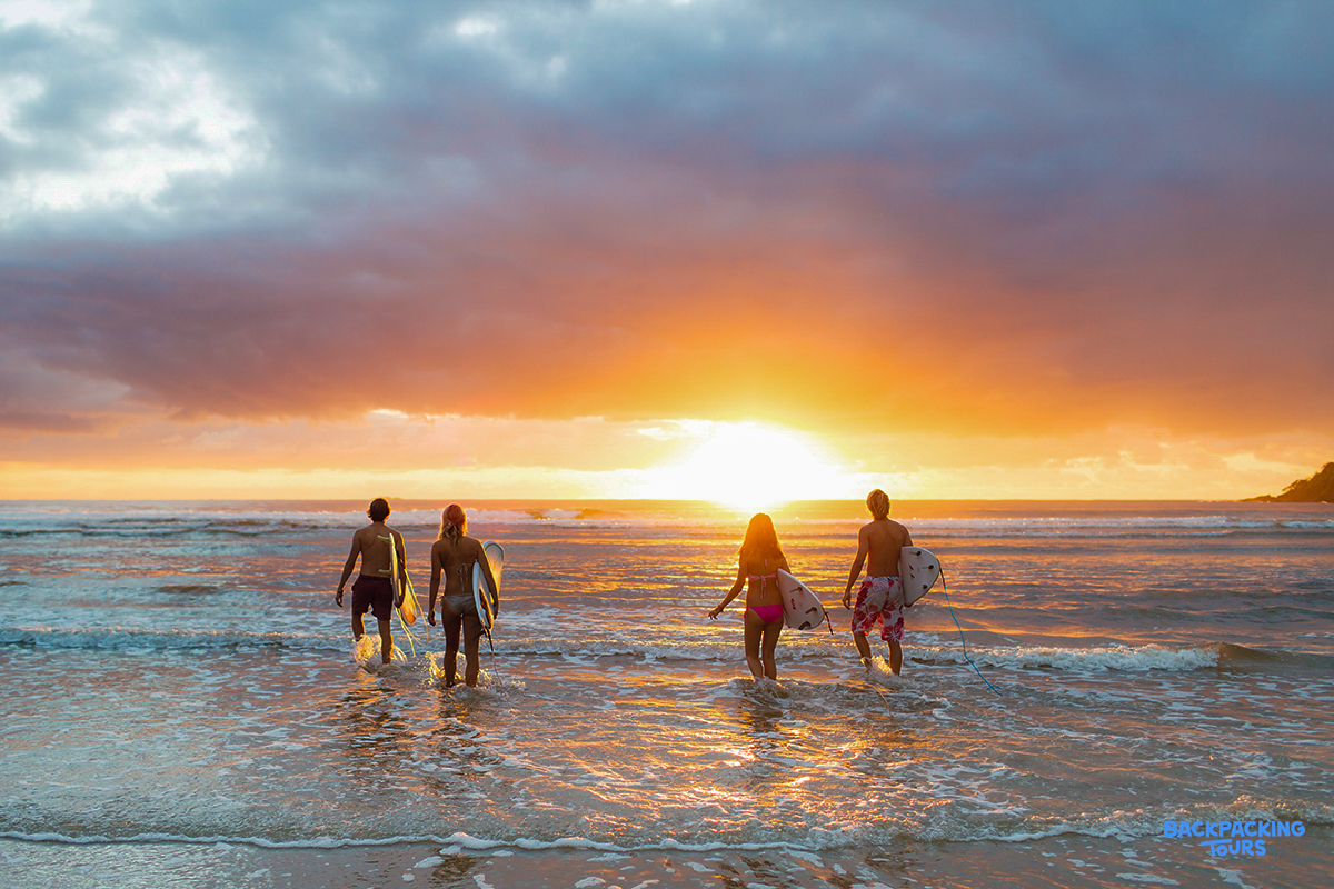 Four friends with surfboards wade into the ocean at Byron Bay during a stunning orange sunset.