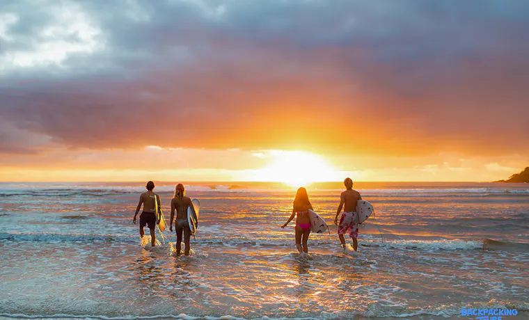 Backpackers carrying surfboards walk into the waves at a vibrant Byron Bay sunset.