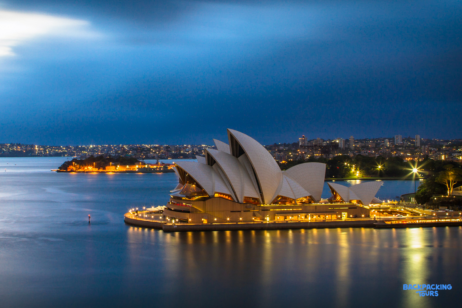 The iconic Sydney Opera House glows brightly at dusk, reflecting on the calm harbor waters.