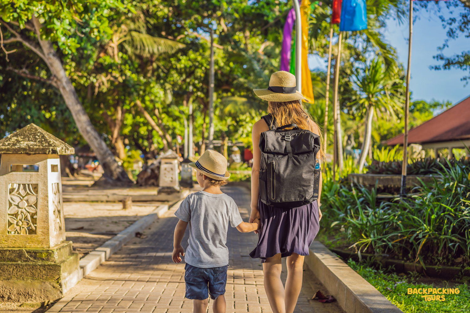 A mother and child walk hand-in-hand along a tropical path with colorful flags in Ubud, Bali.