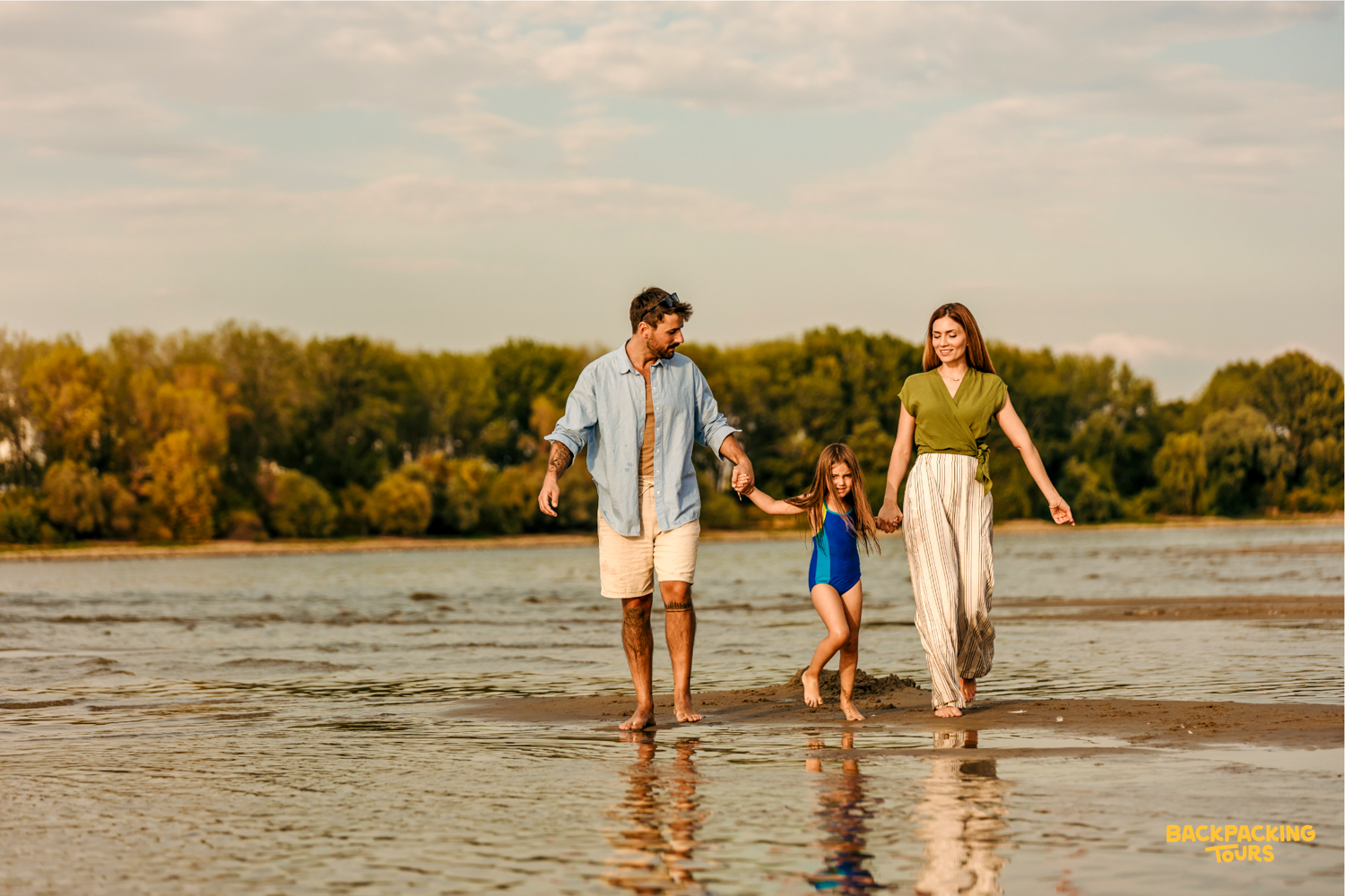 A happy family walks barefoot, holding hands, on a sandbar in shallow water at sunset.