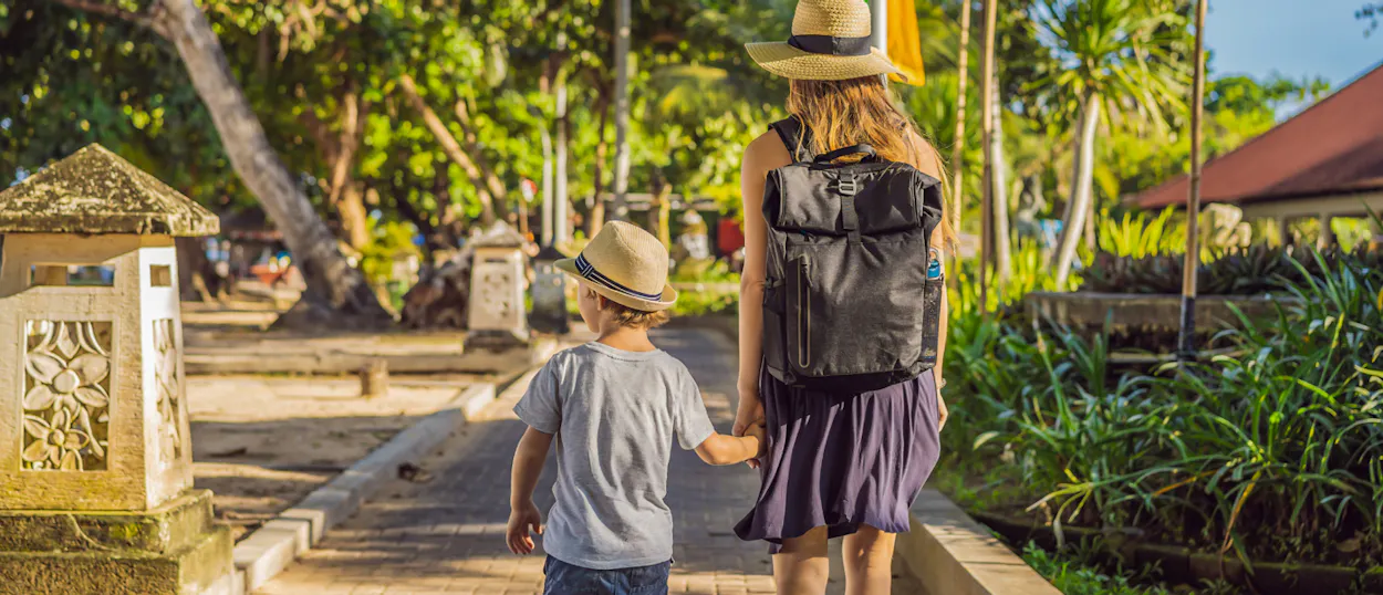A woman and child with hats walk hand-in-hand on a path through a tropical forest in Ubud, Bali.