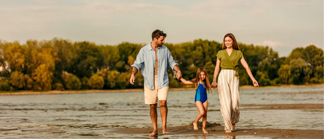 A smiling family holding hands walks barefoot on a wet sandy shore during golden hour.