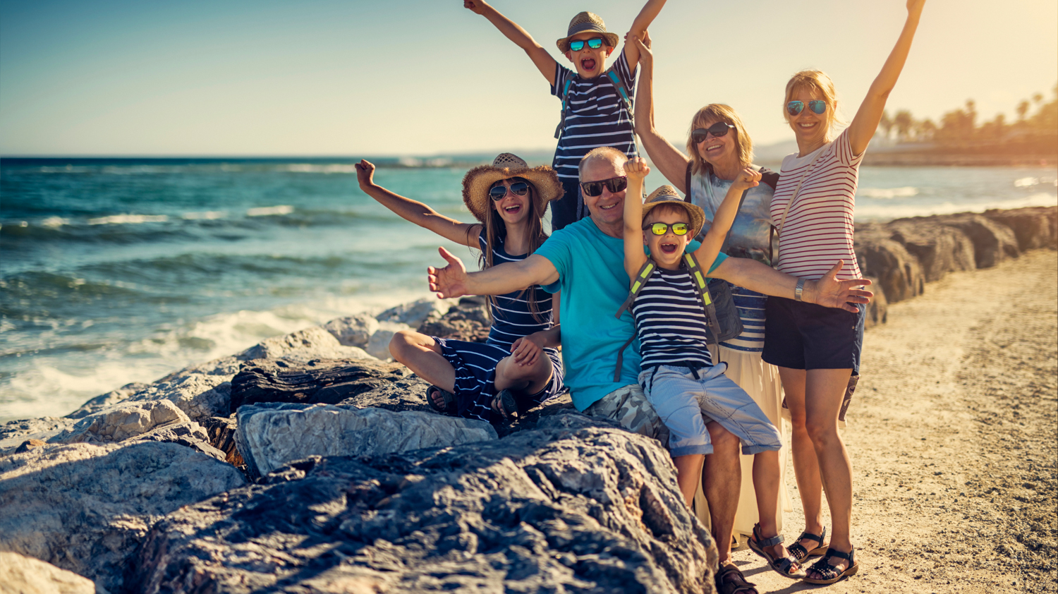 A multi-generational family joyfully poses with arms raised on rocks by the sunny ocean.