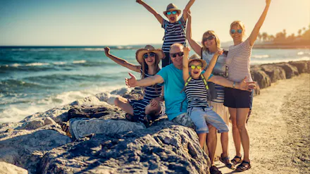 A multi-generational family joyfully poses with arms raised on rocks by the sunny ocean.