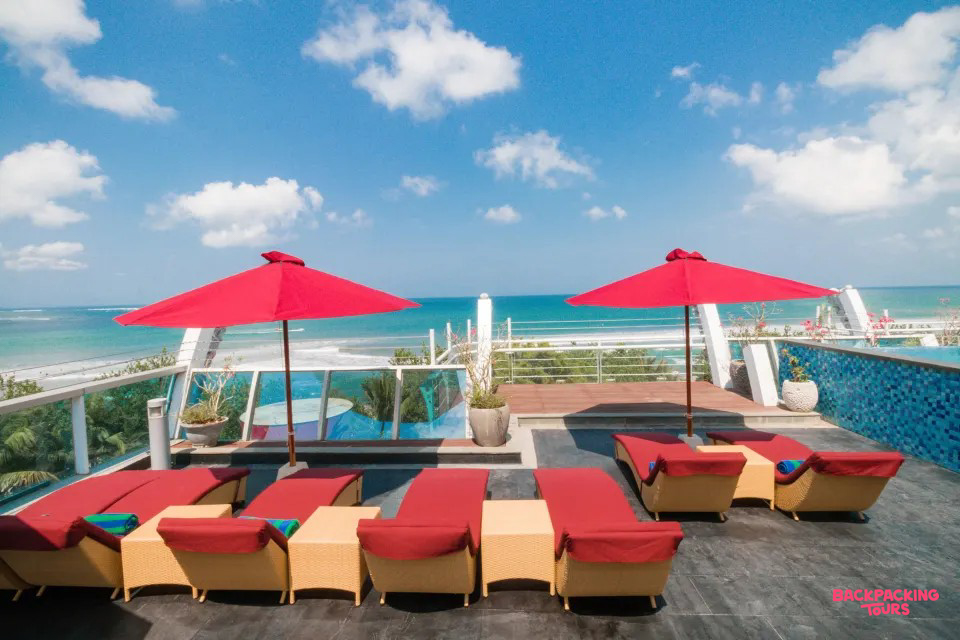 Rooftop terrace with red sun loungers, umbrellas, and a pool overlooking the Kuta beach.