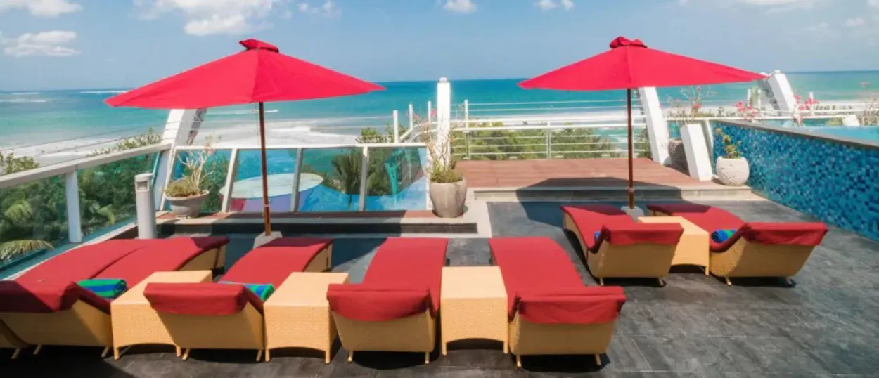 Relaxing rooftop pool deck with red lounge chairs and umbrellas overlooking the Kuta beach and ocean.