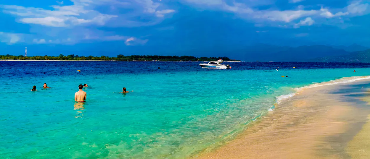 People swim in the clear turquoise waters of Gili Trawangan, with a boat and island in the distance.