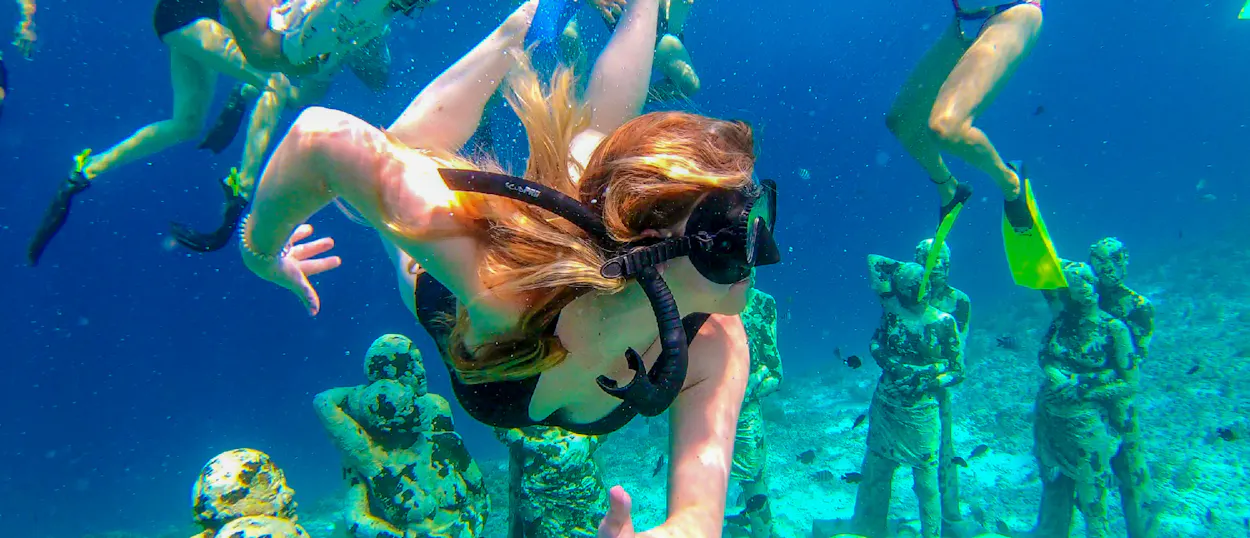 A person with red hair snorkels among underwater statues at Gili Trawangan, surrounded by other swimmers.