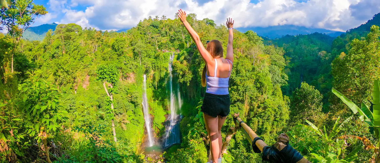 Joyful woman with arms raised, admiring multiple waterfalls cascading through a lush green jungle in Bali.