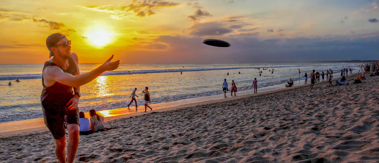 A man throws a frisbee on a vibrant Kuta beach at sunset, with many people enjoying the water.