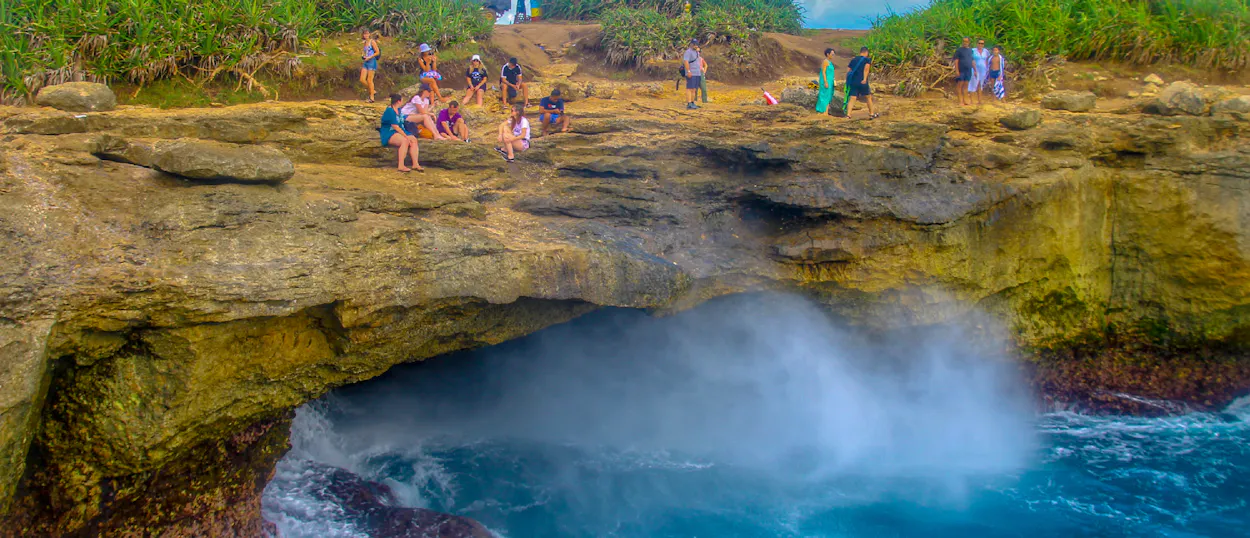 Travelers gather on rocky cliffs watching a powerful wave spray at Devil's Tear, Nusa Lembongan.