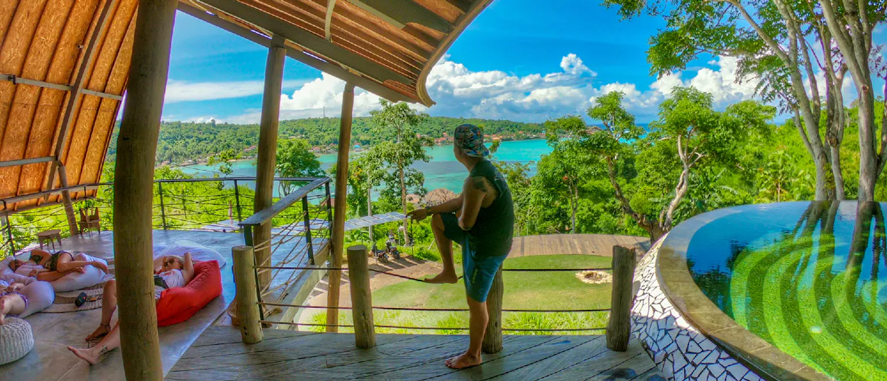 A person stands on a balcony overlooking the vibrant turquoise ocean and lush islands of Nusa Lembongan.