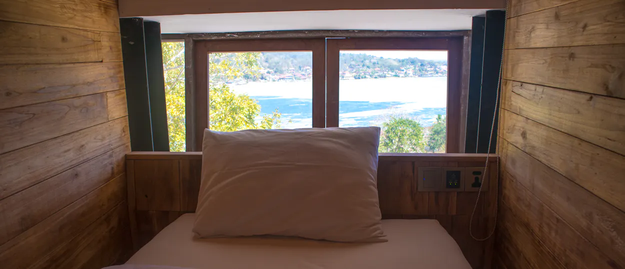 A bed in a wooden room with a window view of the blue ocean and distant islands.