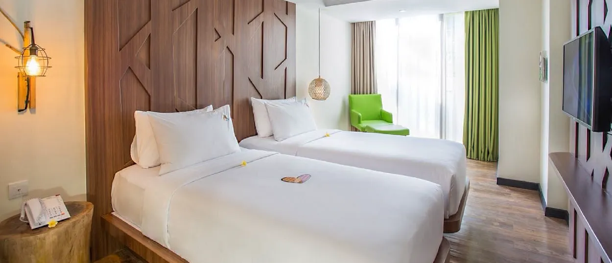A bright, modern hotel room featuring two single beds, a patterned wooden headboard, and a green lounge chair.
