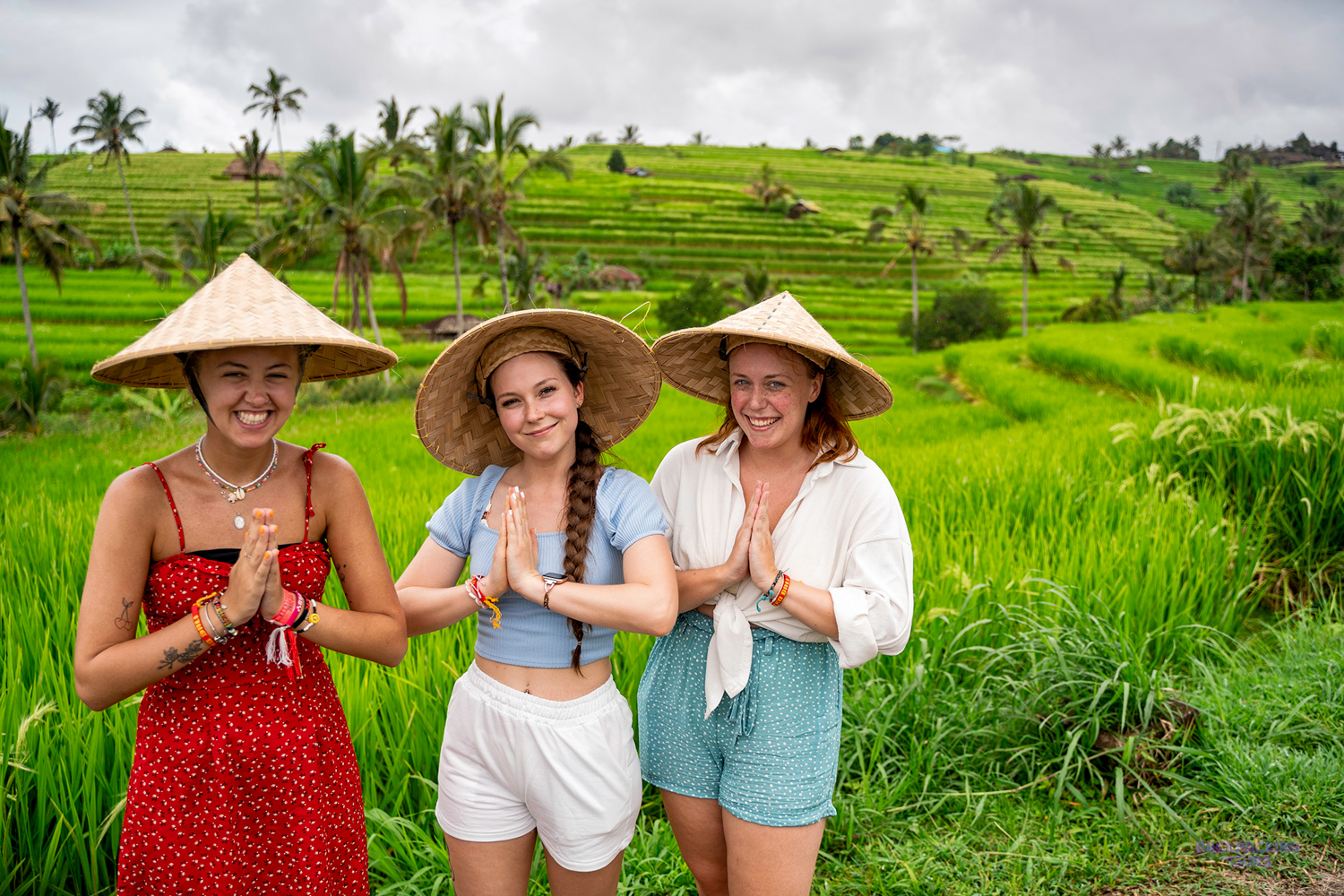 Three happy women in traditional hats pose with Namaste gesture in lush green Bali rice terraces.