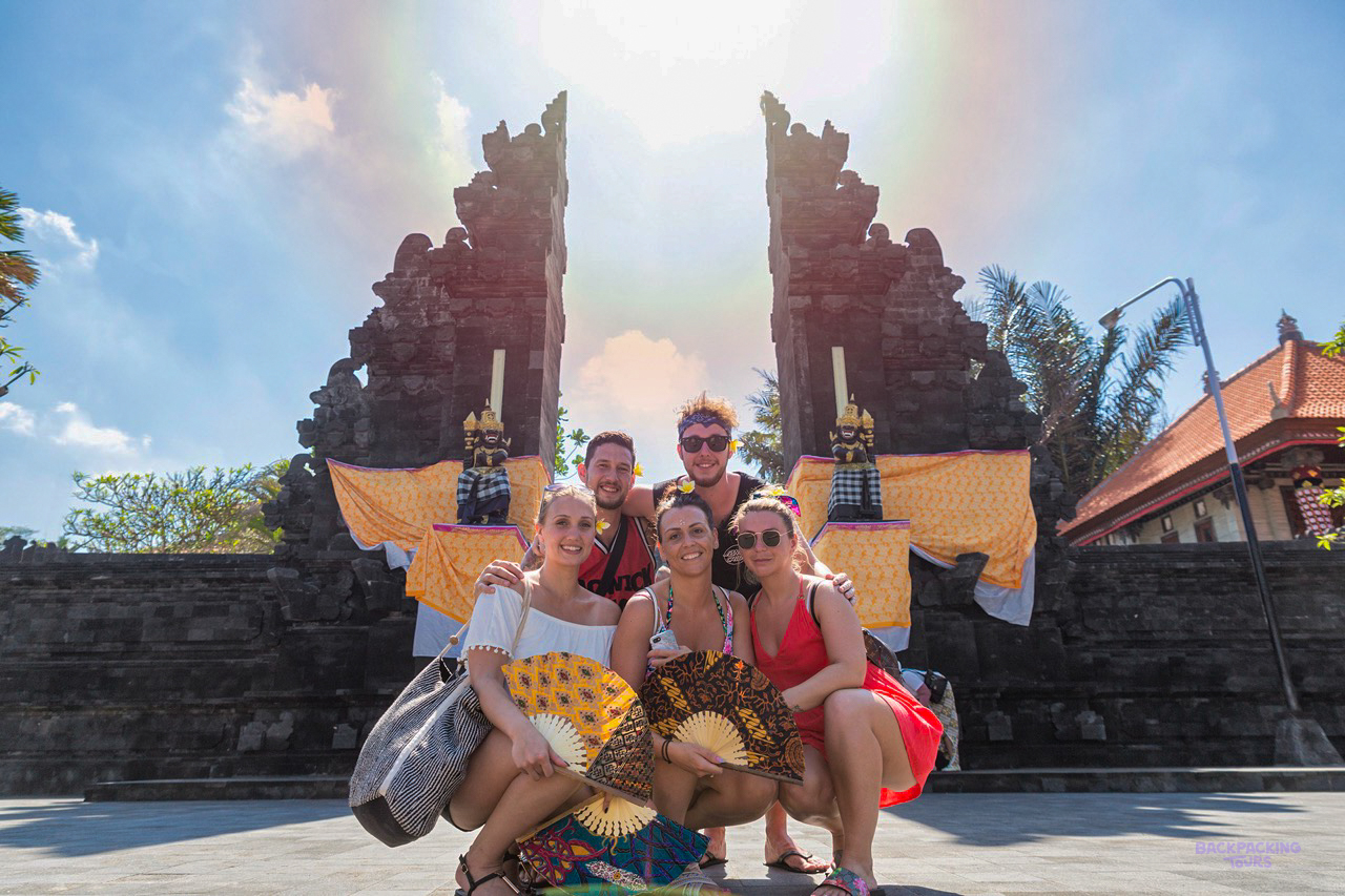 Five happy friends pose in front of a traditional Balinese temple gate on a sunny day.