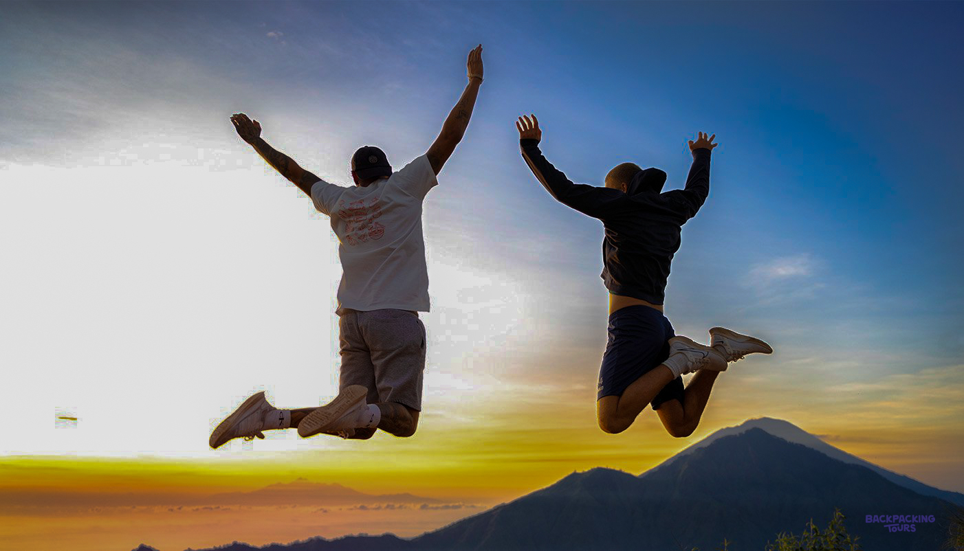 Two joyful travelers jump high against a vibrant sunrise over Mount Batur in Bali.