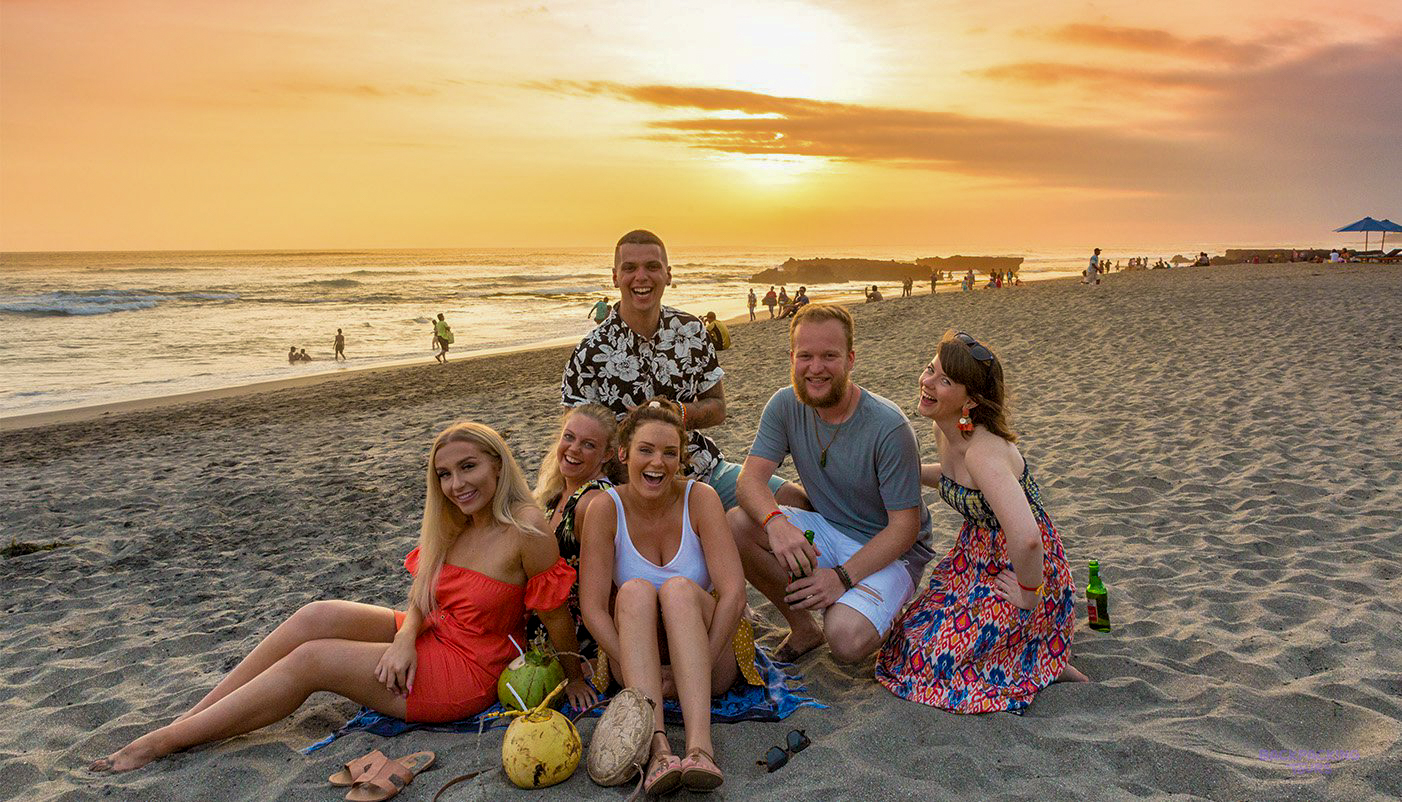 Six friends smiling and relaxing on Canggu beach at sunset, with coconuts on the sand.