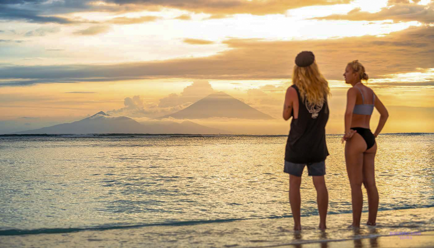 Two travelers watch a golden sunset over the ocean and distant mountains from Gili Trawangan beach.